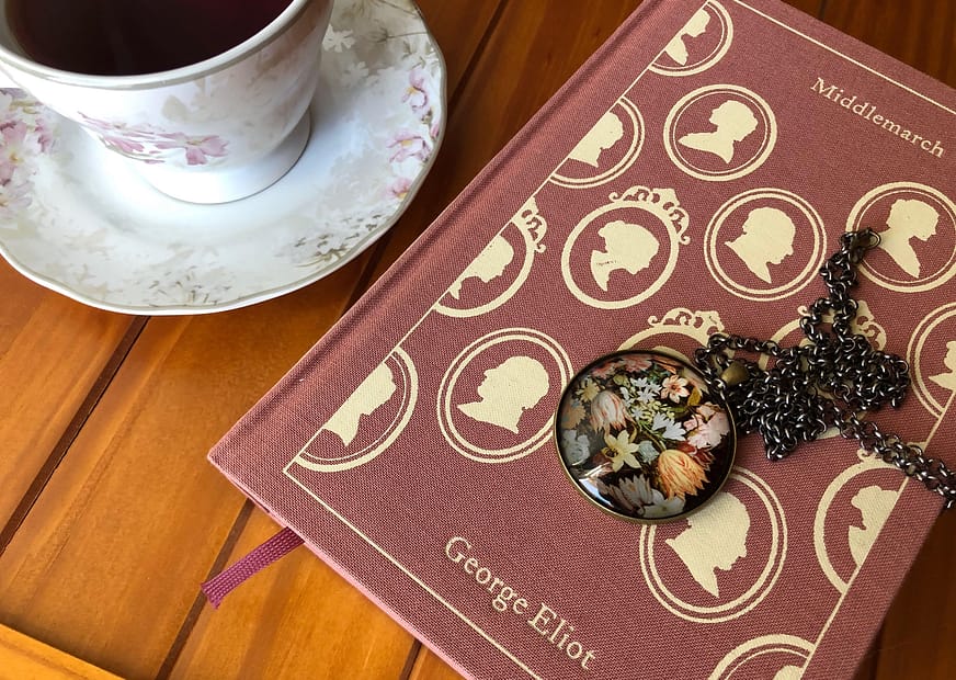 Foto do romance Middlemarch de George Eliot sobre uma superfície de madeira. À esquerda do livro, há uma xícara de chá de porcelana e sobre a capa da obra um cordão com pingente floral.